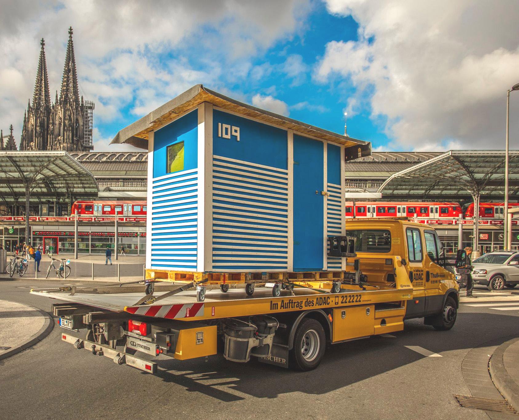 Eine blau-weiße Wohnbox von Little Home auf einem Transporter vor dem Kölner Hauptbahnhof und dem Dom